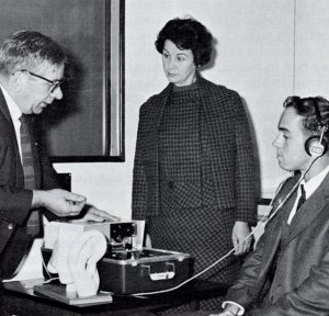 A black and white image of three people testing an audiometer, one man has headphones on his ears while a woman looks on and another man is testing a read-out from the machine.