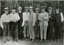 An image of seven men in suits posing in front of a barn door.