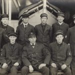 Group portraits of nine men in uniform before a boat.