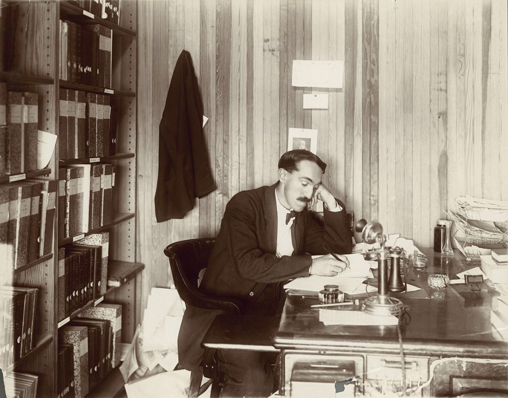 A sepia portrait of a man with a large mustache leaning on his hand at a desk while writing on some papers.