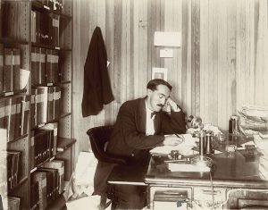 a sepia portrait of a man with a large mustache leaning on his hand at a desk while writing one some papers