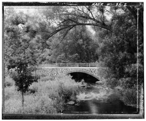 black and white photo of the wolf river going under a bridge in a wooded area with a meadow on the left side