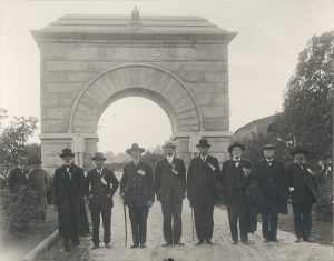 Black and white image showing a group of men in suits standing in front of the Camp Randal arch with Old Abe on top of the monument