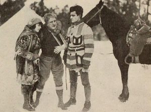 a film still from the 1919 movie, A wilderness trail, showing three people standing before a horse in the woods. One man is wearing a short point blanket coat.