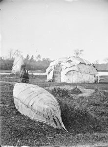 A black and white image of a Ho Chunk person leaning on a beached canoe with a wigwam and tipi in the background