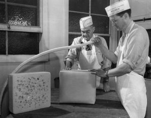 A black and white image of two men cutting Swiss cheese