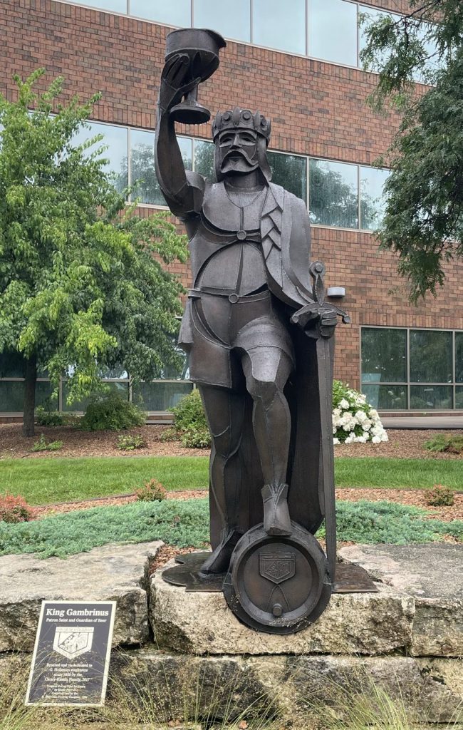 A metal statue of a king with a sword in one hand, the other hand raises a chalice into the air, he stands with one leg bent, foot resting on a barrel.