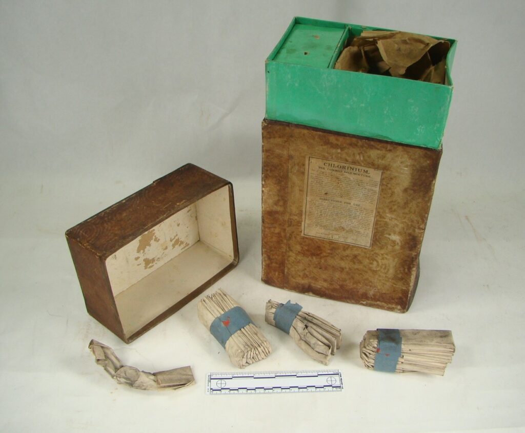 Weathered cardboard box with text label, the lid is removed revealing a aqua colored interior. The contents, paper-wrapped chlorinium packets are bound in blue tape.