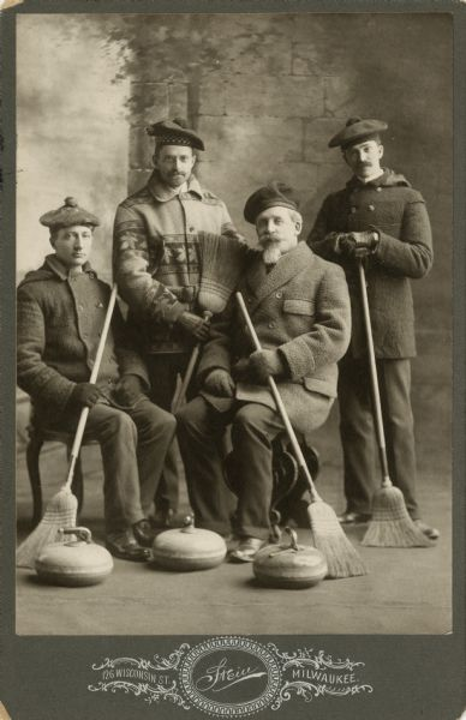 A sepia-tone studio photograph of four men in winter coats and tams holding straw brooms posed behind three curling stones.