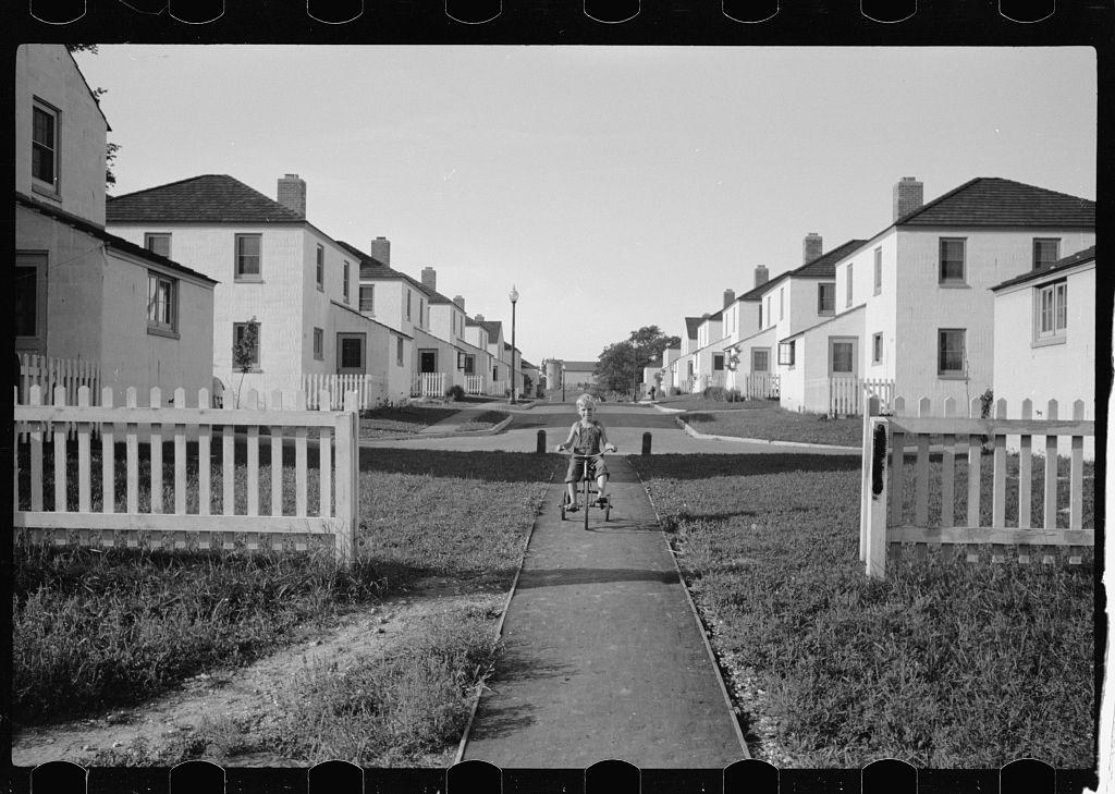 A black and white photograph of a boy riding a tricycle on a sidewalk between identical houses.