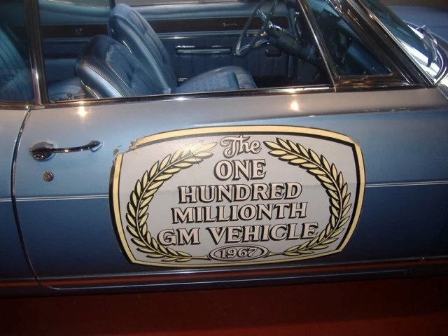 Close-up image of a white sticker with gold lettering affixed to a car door that reads, "The One Hundred Millionth GM Vehicle"