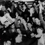 A crowd of people cheer before an elevated speaker. The newspaper the speaker holds has the headline "Strike is Settled."