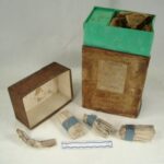 Weathered cardboard box with text label, the lid is removed revealing a aqua colored interior. The contents, paper-wrapped chlorinium packets are bound in blue tape.