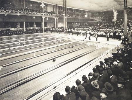 Photograph of a large bowling hall with a dozen lanes with men bowling. Spectators look on from grandstands and balconies.