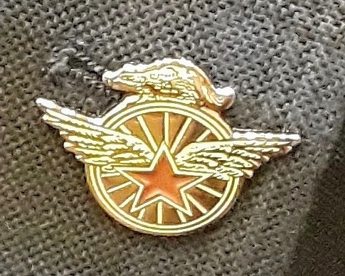 A brass and enamel pin featuring a winged star imposed over a spoked bicycle wheel, atop of which stands the figure of a badger.
