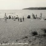 A black-and-white image of figures on a sandy beach and bathing in shallow water.