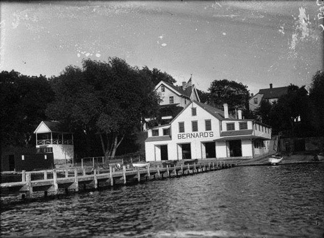 A black-and-white photograph of a boathouse and long pier.