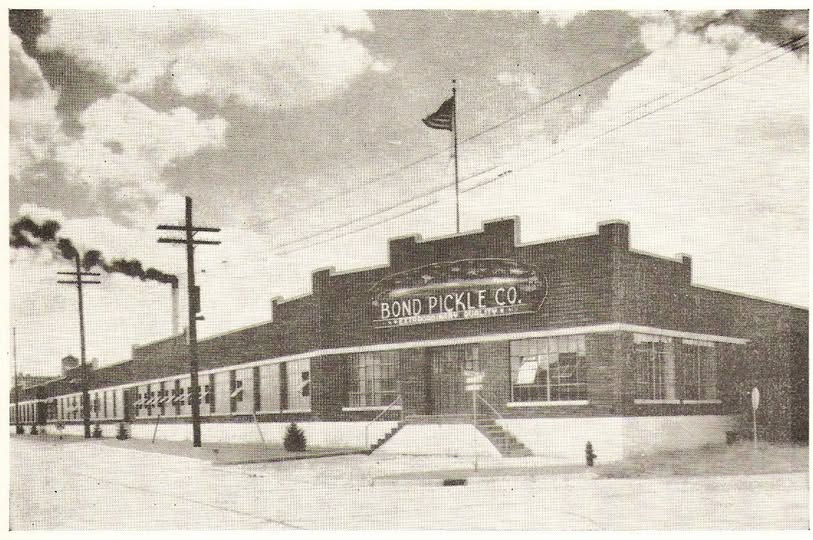 Exterior photograph of a large one-story brick factory building. A sign on the front of the building reads, 'Bond Pickle Co.'