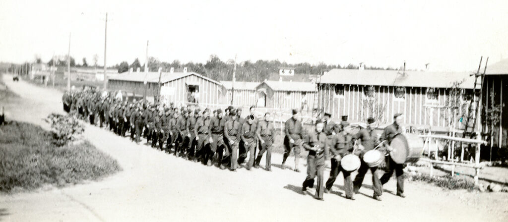 Black and white photograph of men in military uniforms marching in front of temporary buildings.
