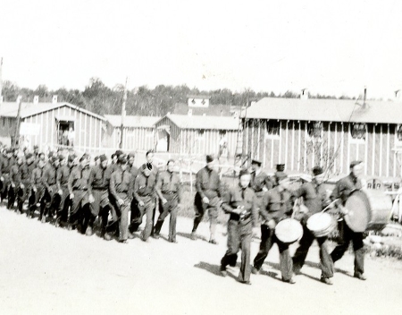 Black and white photograph of men in military uniforms marching in front of temporary buildings.