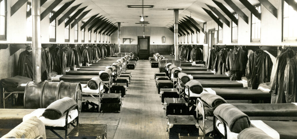 Interior view of a barracks with two rows of neat beds, each with rolled blanket and trunk.