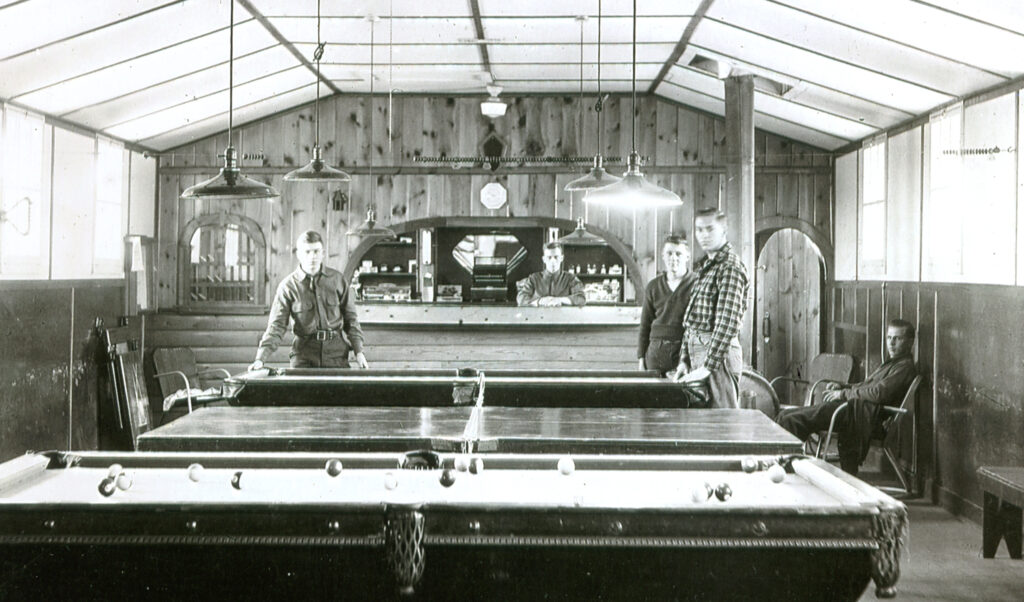 Interior view of a recreation hall with billiards and table tennis tables. Three men pose next to one of the game tables. In the background, another man leans over a commissary counter.