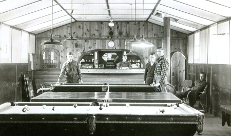 Interior view of a recreation hall with billiards and table tennis tables. Three men pose next to one of the game tables. In the background, another man leans over a commissary counter.