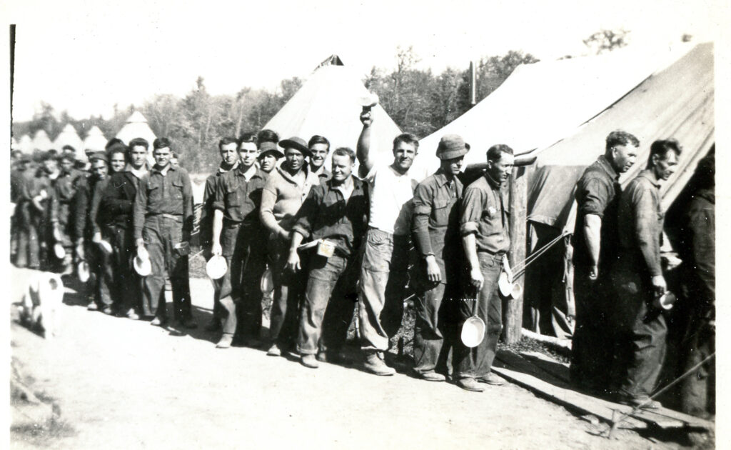 Men in work clothes holding bowls wait in line before a large canvas tent.