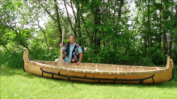 A man poses with a oar and a birchbark canoe.