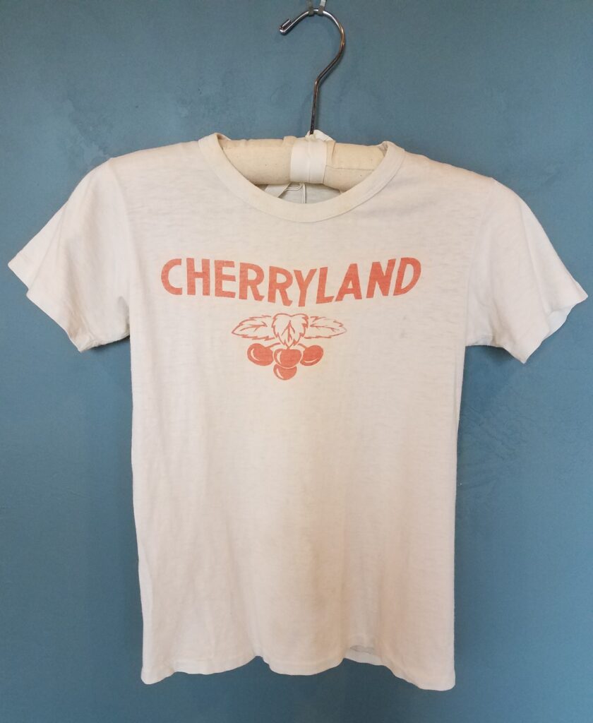 A white T-shirt on a hanger with red printing of a cluster of cherries and the word “Cherryland”