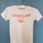 A white T-shirt on a hanger with red printing of a cluster of cherries and the word “Cherryland”