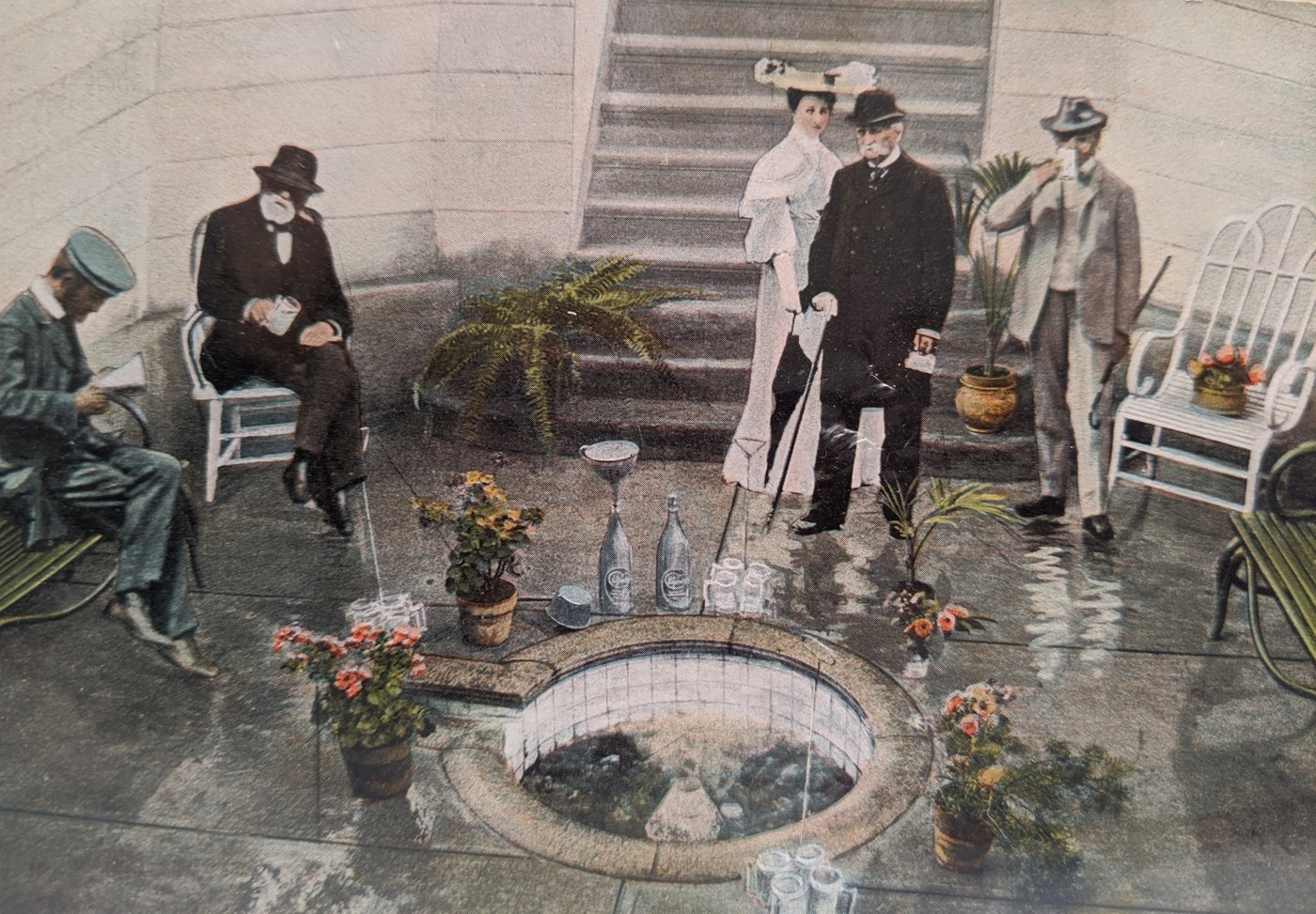 Colored photographic postcard showing men and women in finery around a small spring. An attendant sits by, and there are cups and bottles around the spring well.