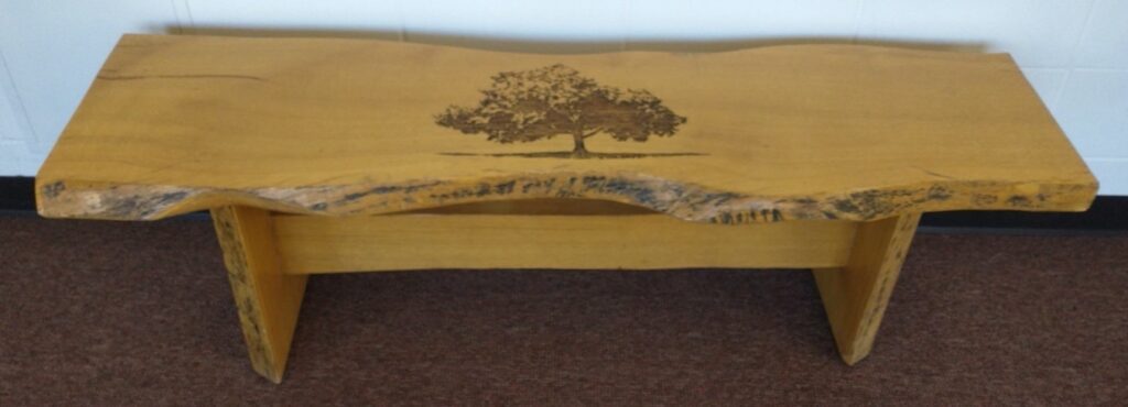 A live-edge wooden bench with a laser engraving of an oak tree on the seat.