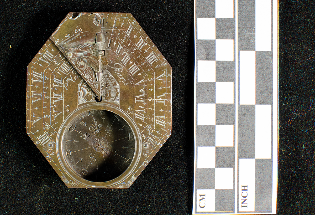 An octagonal, engraved brass sundial and compass. A ruler indicates that it is approximately 8 centimeters (3 inches) long.