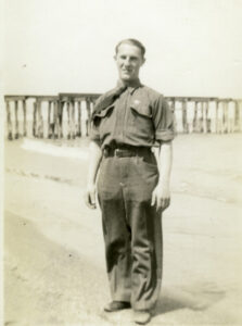 A uniformed man stands posed on a beach.