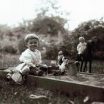 A black-and-white photograph of a young boy with dolls, stuffed animals, and a tea kettle in a sandbox.