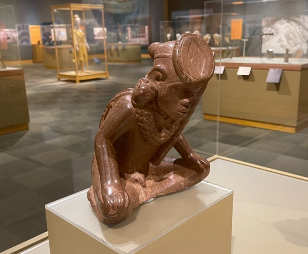 Red bauxite effigy pipe sculpture of a male figure sitting cross-legged with his hands resting on his knees wearing maskettes as ear jewelry.