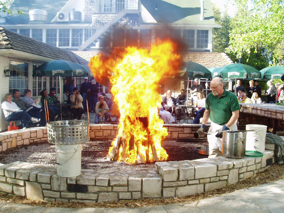 A man in a large stone-lined ring walks around a large open fire. The accoutrement of a fishboil, a boil pot, and strainer basket, plastic buckets, and a two-sided spork are at hand. A crowd of people seated at tables watch on.