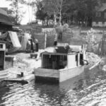 A black-and-white photograph of two small wooden boats docked next to a small house. Two men work on fishing nets.