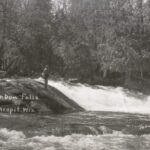 A black-and-white photograph of a man standing on a large rock in the middle of a river near a falls, fishing.