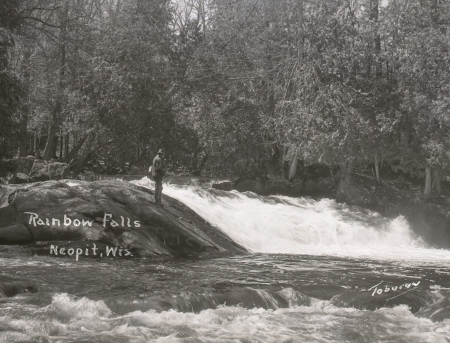A black-and-white photograph of a man standing on a large rock in the middle of a river near a falls, fishing.