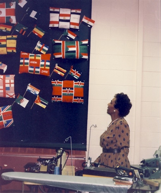 Earlene Fuller stands in front of a classroom gesturing at a large board upon which are pinned Kente cloth patterned textiles.
