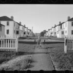 A black and white photograph of a boy riding a tricycle on a sidewalk between identical houses.
