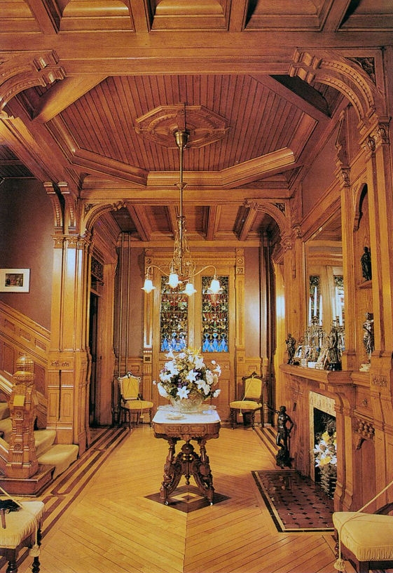 Photograph of the interior of the foyer of the Hearthstone house. The entryway features detailed wood ornamentation, and at the center of the room is a four-light electric chandelier.