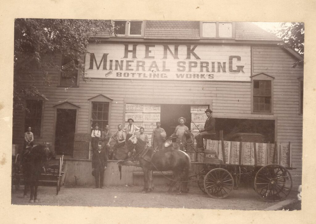Photograph of a horse and cart and a group of men posed before a building with a large sign that reads “Henk Mineral Spring Bottling Work’s”