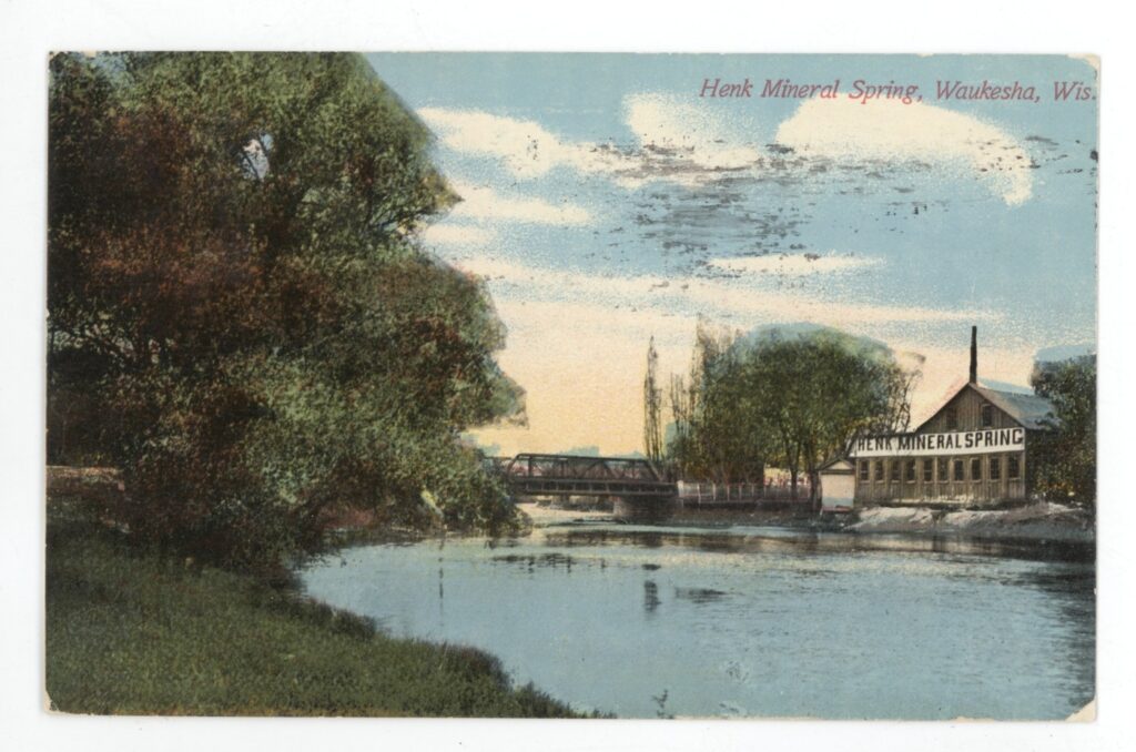 Colored photographic postcard of a view of the Henk Mineral Spring Bottling Company factory building across a pond.