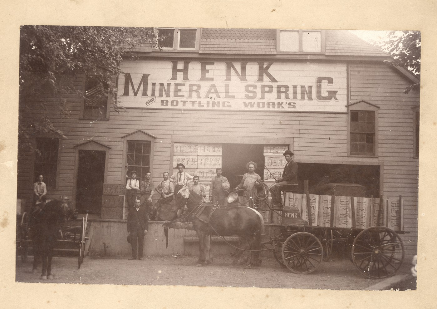 Photograph of a horse and cart and a group of men posed before a building with a large sign that reads “Henk Mineral Spring Bottling Work’s”