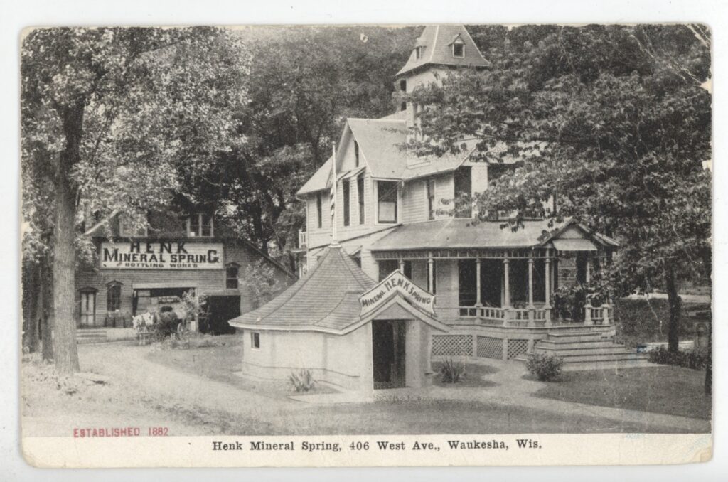 Photographic postcard of the Henk Mineral Spring, including the springhouse, a large home, and the bottling works in the background.