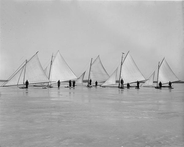 A black and white photograph if five ice boats on a frozen lake.