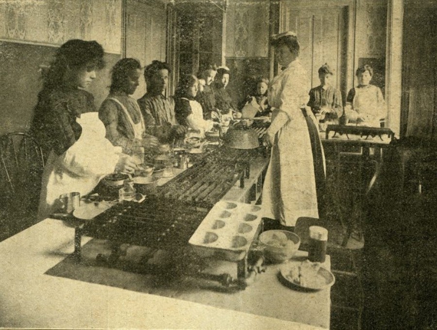 A woman stands before, and teaching, a group of women who work over table-top grills and cook pots.